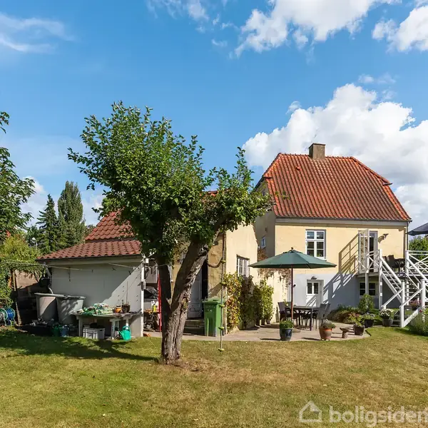 Et gult hus med rødt tag står i en have. Huset har en trappe og terrasse til højre. Haven er græsbeklædt med et centralt træ og omgivet af buske og træer.