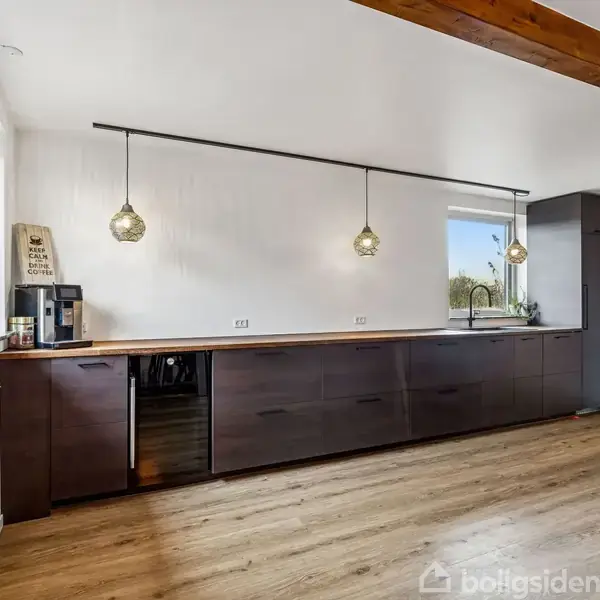 Et køkken med mørke skabe strækker sig langs en væg, oplyst af to hængelamper. Til venstre står en kaffemaskine ved siden af en grøn plante ved vinduet.