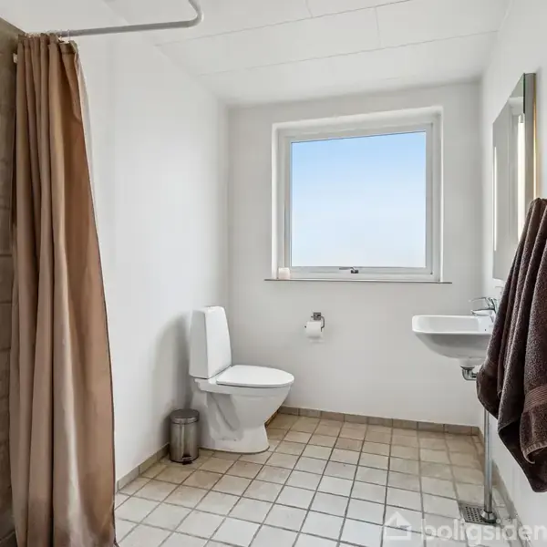 Toilet med cisterne står i et badeværelse. Et bruseforhæng hænger til venstre, mens et spejl og en håndvask med håndklæder hænger på væggen til højre.