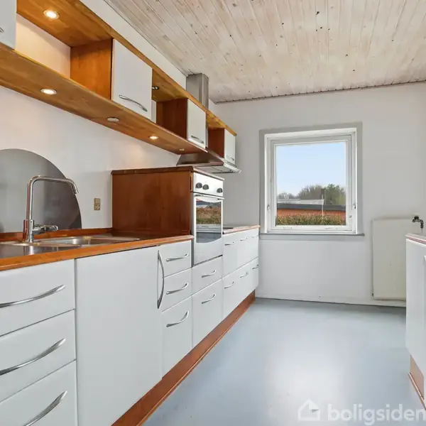 Køkken med hvide skabe og skuffer, træbordplade, indbygget ovn, og vask. Hvide vægge og loft, gråt gulv. Et vindue med udsigt til grønt landskab.