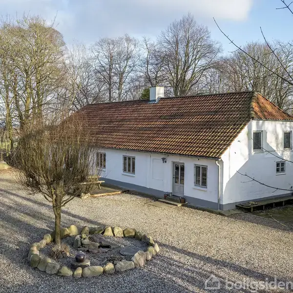 Hvidmalet hus med rødt tegltag står stille på en grusindkørsel omgivet af træer. Foran huset er en cirkulær stensætning med et træ i midten.
