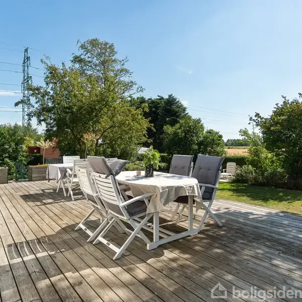 En udendørs terrasse med spisebord, omgivet af grønne træer og buske, under en klar blå himmel. Bordet har en dug og stole står omkring det.