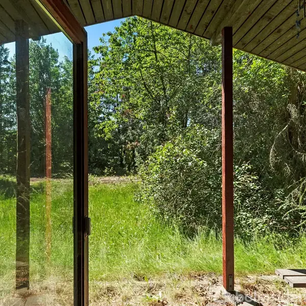 Glasdør åbner ud til en frodig have med græs og træer i baggrunden. Træterrasse og skovområde omkranser området i en naturskøn, uforstyrret beliggenhed.