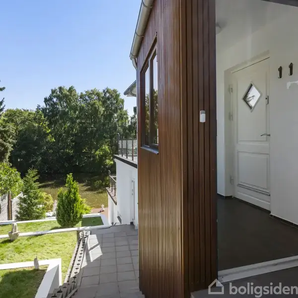 En hvid dør står åben ind til et hus med trægavl. Udenfor er en flisebelagt terrasse med udsigt til en grøn have med træer og plæner i baggrunden.