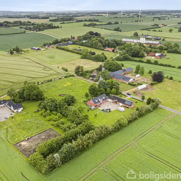 Luftfoto af landdistrikt viser gårde omgivet af grønt landskab med marker og træer. I forgrunden ses en gård med flere bygninger og en indkørsel med parkerede biler.