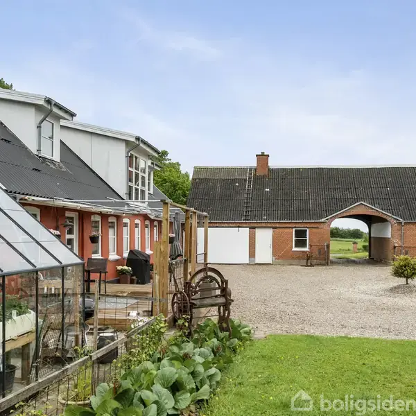 Væksthus står ved siden af en rød murstensbygning med sorte tage. I baggrunden ses en stor lade i landdistriktsområde. Fremsiden har grus og en lille busk.