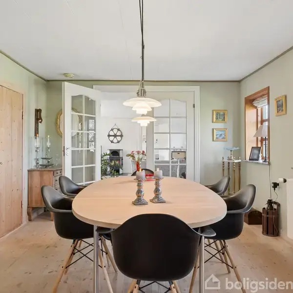 Rundt spisebord med sorte stole står under en lampe i et lyst rum. Væggene har kunstværker, og der er trægulv. Vinduer og en dør udsigt til et tilstødende værelse.