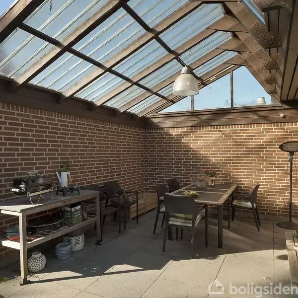En udendørs terrasse afskærmet med glastag, indeholder spisebord og stole. Murede vægge omkranser området, der har dagslys fra ovennævnte glastag. En grillstation står langs muren.