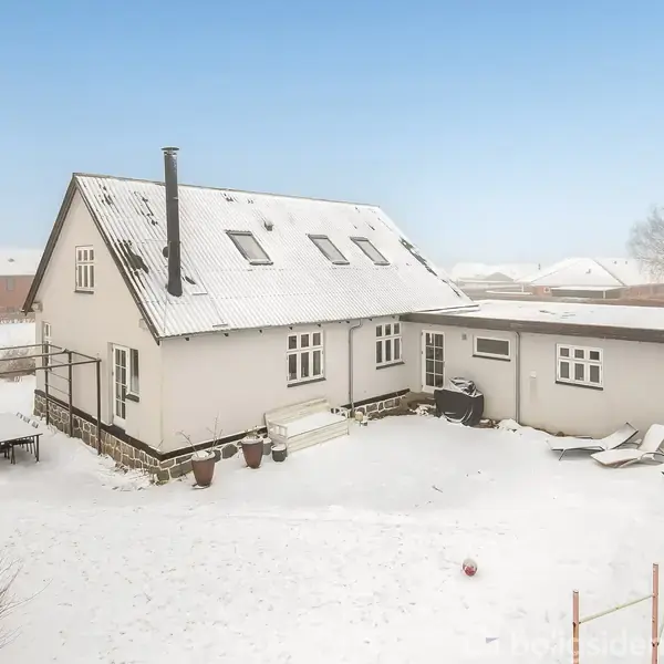 Et hvidt hus står i en snedækket have med solstole og et bord med stole. Himlen er klar, og der er et hegn i baggrunden.
