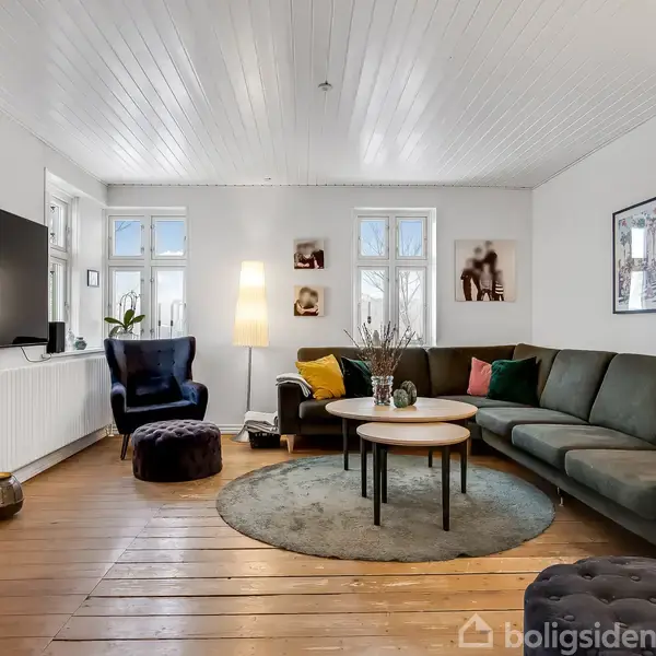 Stue: Stor grå sofa står i hjørnet. En blå lænestol ved siden af en læselampe. Rundt tæppe og sofabord i midten. Fladskærm på væggen. Vinduer med lysindfald.