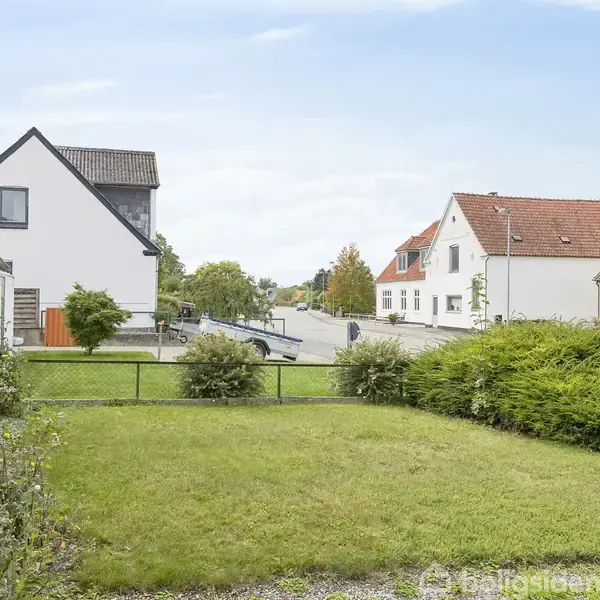 Græsplæne omgivet af buske og hæk i forgrunden. Baggrunden viser en række huse i forskellige arkitektoniske stilarter langs en rolig vej. Himlen er delvist skyet.