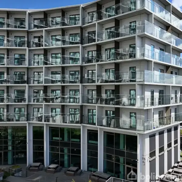 Bygning med moderne lejligheder; mange altaner med glasværn. Nederste etage har store vinduer. Omkring er der et grønt område og flere stier under en delvist skyet blå himmel.