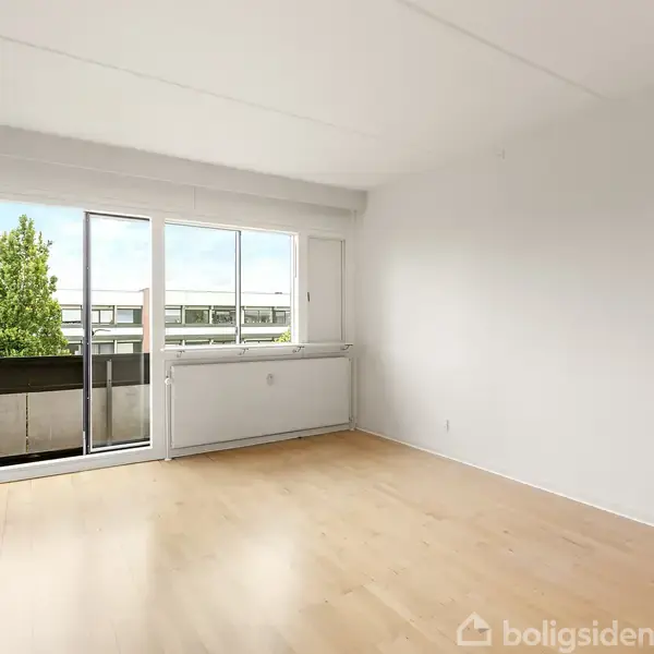 Et lyst rum med trægulv og lyse vægge, der har en altandør og vindue mod en balkon med udsigt over et grønt område og huse i baggrunden.