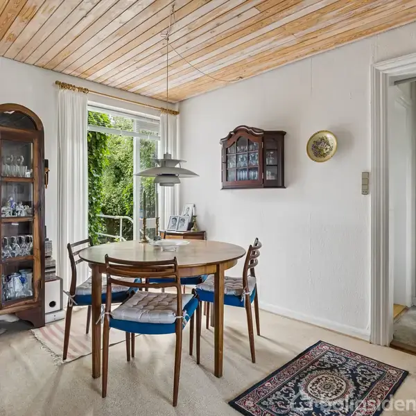 Et rundt spisebord med fire stole står i et lyst rum. Ved siden af, et vitrineskab med glas. En dør fører ud til en terrasse med grønt udenfor. En vægklokke hænger ved siden af en mindre skænk.