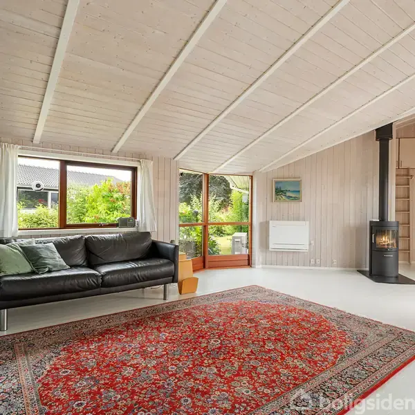 Sort sofa står ved vindue; stor rød orientalsk tæppe på gulvet. Trævægge og loft med træplader; brændeovn i hjørnet; grøn have udenfor; lys og rummelig stue.