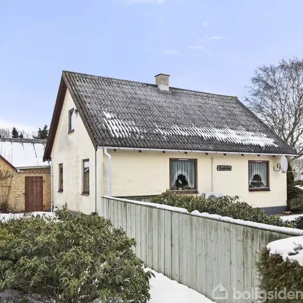 Et lille, hvidt hus med et skifertag er dækket af let sne. Foran huset er der en grøn hæk og en snebelagt vej. En nabobygning ses til venstre.