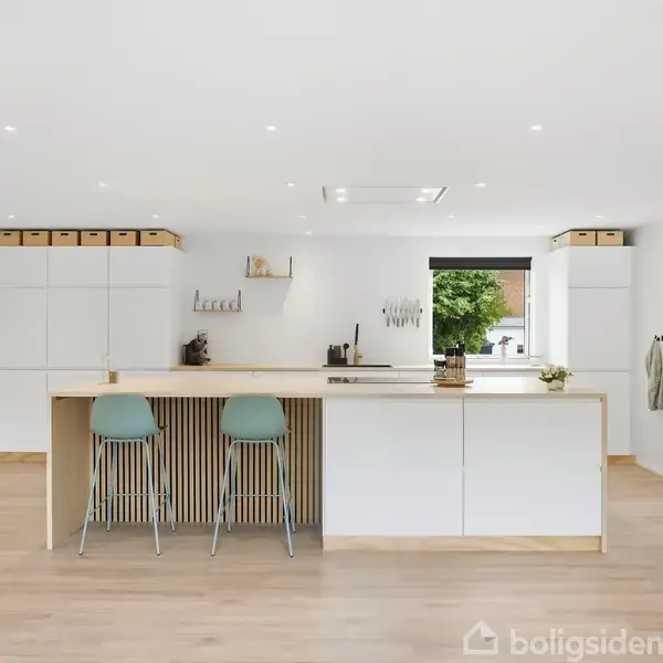 Et moderne køkken med en stor ø i midten, udstyret med to barstole. Rummet er lyst, med indbygget opbevaring, vindue og integreret ovn i et minimalistisk design.