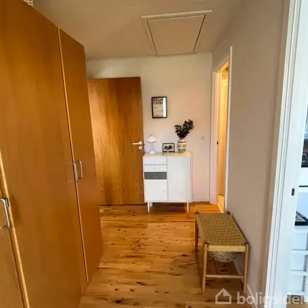 Skabe står langs en gang med trægulv. En hvid kommode med dekorationer står for enden. Der er døre til højre og en bænk nær åbningen til et værelse.