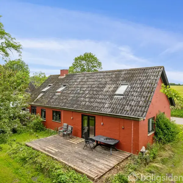 Et rødt hus med skråtag og flere vinduer står på landet. En træterrasse med havemøbler er foran huset. Omgivet af frodig grøn vegetation, åbne marker og en blå himmel.