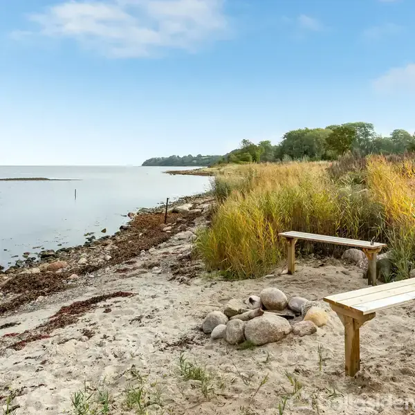 To bænke står ved en sandstrand med sten og græs nær vandet. Baggrunden viser en kystlinje med træer i horisonten under en blå himmel.