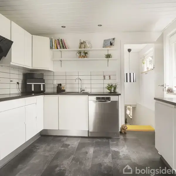 Et moderne køkken med hvide skabe og en grå flisebelagt gulv. En opvaskemaskine og sort ovn er indbygget. Ovenfor er hylder med bøger og planter. En stor vindue til venstre.