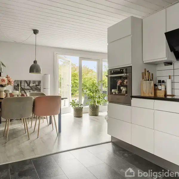 Et moderne køkken med hvide skabe og en ovn, åben til en spisestue med pastelfarvede stole omkring et bord, blomster og planter foran store vinduer.