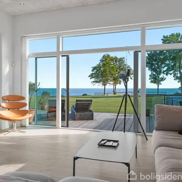 En moderne stue har store vinduer med udsigt til en terrasse, græsplæne og havet. En brun lænestol og en lys sofa dominerer rummet, der er oplyst af naturligt lys.