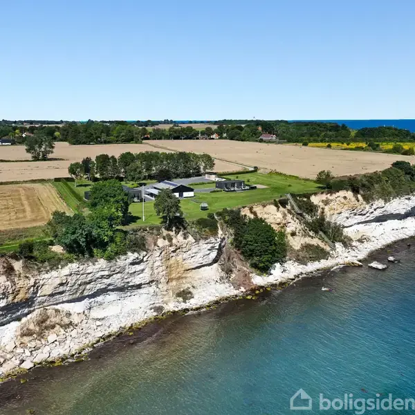 En moderne bolig ligger på en klippekyst omkranset af marker og træer. Kysten strækker sig langs en klar, blå havoverflade, under en klar, blå himmel.