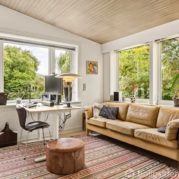 Stue med lysebrun sofa og to puder står foran store vinduer. Et skrivebord med stol, computer og lampe står til venstre. Grønne planter udenfor giver naturligt lysindfald.