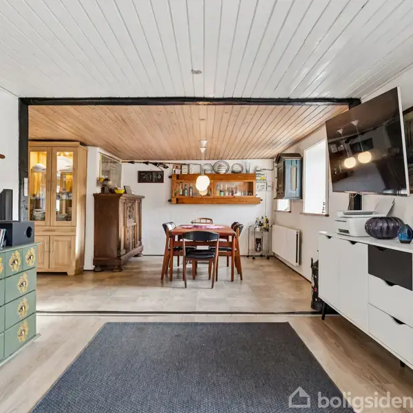 Kommode med grønt mønster står i stue med hvide vægge. TV hænger over hvid skænk til højre. Baggrund viser spisebord med stole foran vitrineskab.