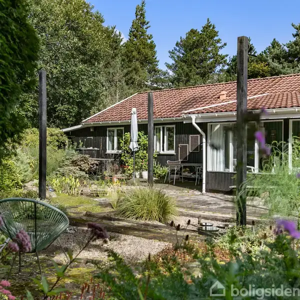 Et hus med rødt tegltag og store vinduer. Omkranset af en frodig have med forskellige planter, træer og en terrasse i forgrunden. Skyfri blå himmel i baggrunden.