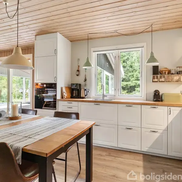 Køkkenbord med fire stole er dækket med en dug og blomster i en vase. Det åbne køkken har hvide skabe, træloft og store vinduer, der lukker naturligt lys ind.