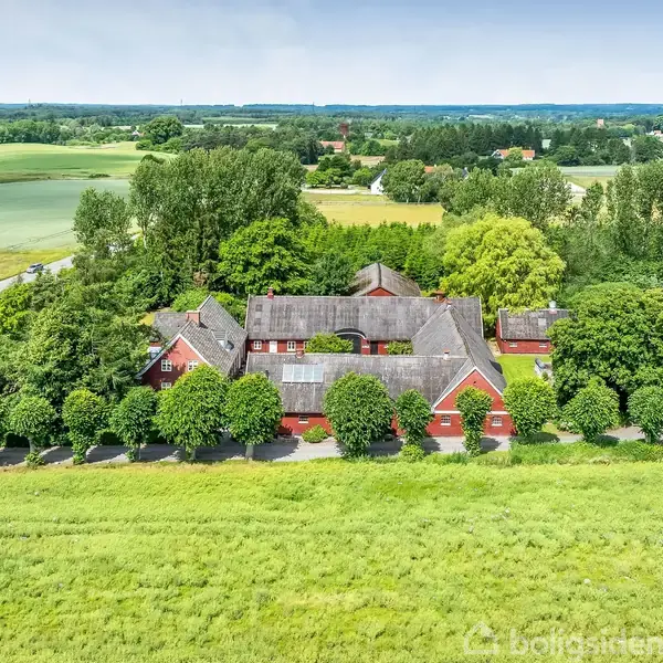 En rød murstensbygning omgivet af tætte træer. Bygningen ligger i et frodigt, landligt område med åbne marker og skove i baggrunden.
