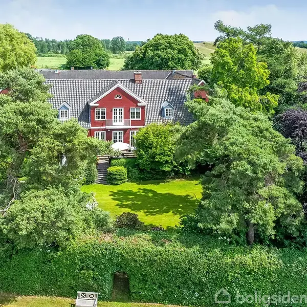 Rød murstensvilla omgivet af frodig have med store træer, beliggende i et landskab med marker og skove i baggrunden under en klar blå himmel.