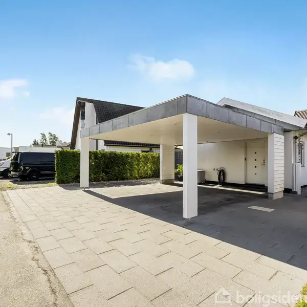 En carport med hvidmalede søjler og tag, står foran et moderne hus. En bred indkørsel fører til en vej med biler og grønne buske til venstre og en sort postkasse markeret "10".