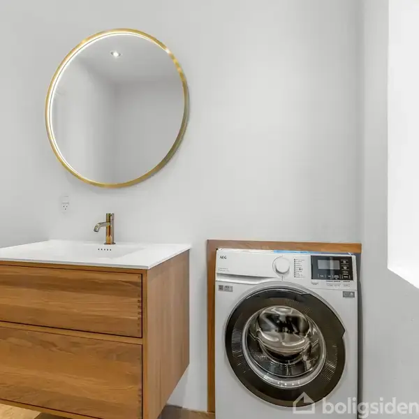 Spejl hænger over et trækonsolbord med vask, guldfarvet vandhane; ved siden står en vaskemaskine. Rummet er lyst og har et vindue til højre.