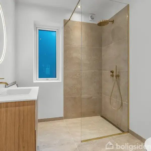 Et moderne badeværelse med brusekabine i glas og guld, en trævask med spejl over. Vindue med blåtonet glas i baggrunden. Enkel og lys indretning.