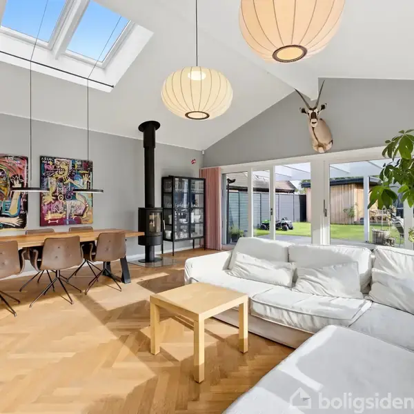 Stue med hvid sofa og sofabord nær glasfacade. Spisebord med seks stole og kunst på væggen. Ovenlys, lofthængte lamper, brændeovn og udsigt til have.
