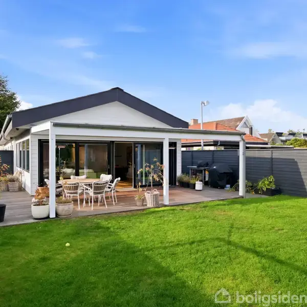 En moderne villa med fladt tag har en terrasse med spisebord og parasol. Terrassen er omgivet af en velplejet græsplæne og indhegnet af et gråt plankeværk.