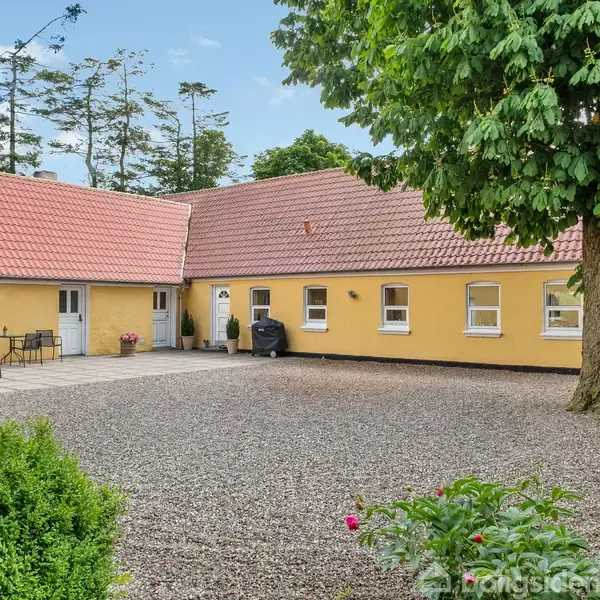 Gult hus med rødt tegltag danner en gårdsplads belagt med grus. Der er en terrasse med havemøbler og urtepotter. Omgivet af grønne træer og buske.