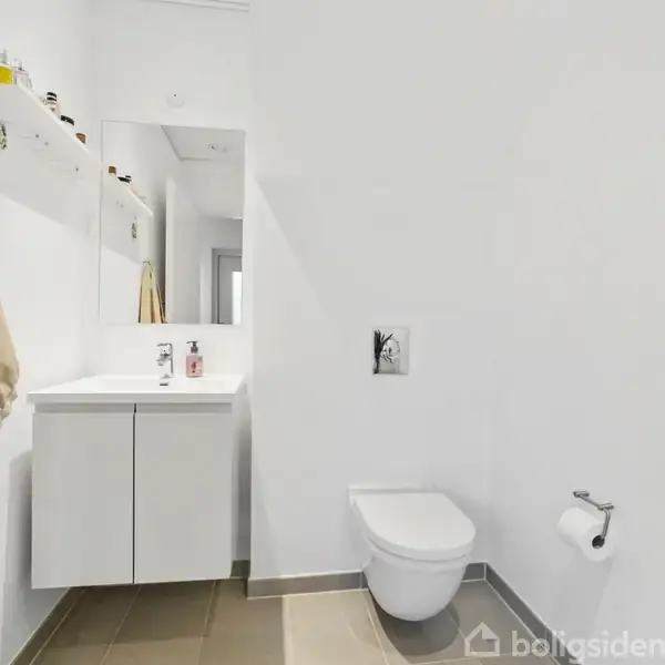 Håndvask med skab under og spejl over hænger på en hvid væg. Ved siden af er et væghængt toilet med toiletpapirholder. Et håndklæde hænger på knop. Flisegulv og hvidt interiør.