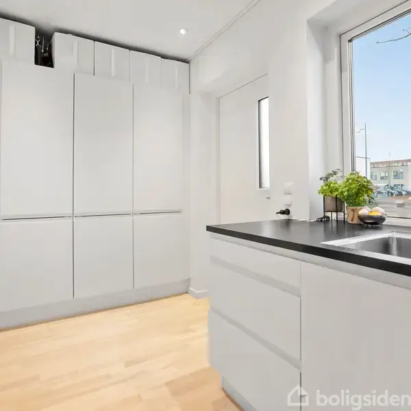 Et lyst køkken med lyse træplanker, hvor en vask er indbygget I en sort bordplade ved vinduet, overskabe i hvid, og håndklæder hænger på væggen ved døren.