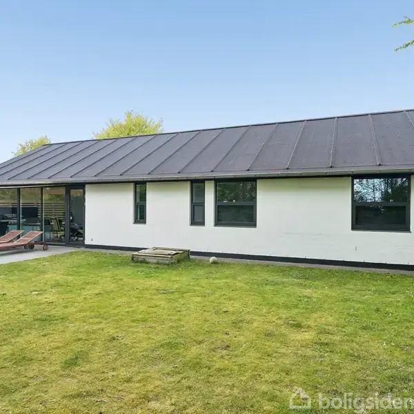 En enkel, hvid etplanshus med sort tag ligger i en velholdt græsplænehave. Ved siden af huset er der en lille terrasse med havemøbler og en grill under en klar blå himmel.