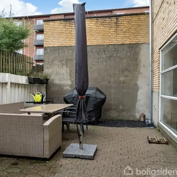 En lukket terrasse med en beige sofa og et bord. En sammenklappet parasol står midt på flisebelægningen. Bagved ses en grill og en høj træhegn, omgivet af murstensbygninger.