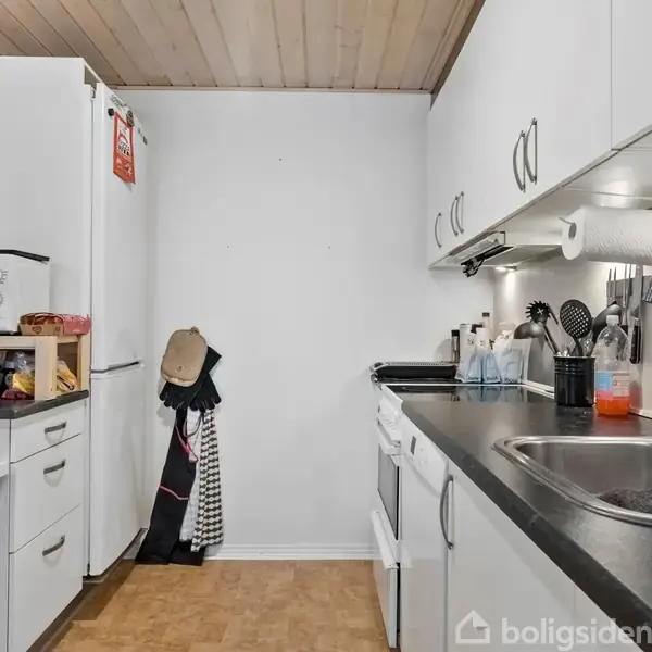 Køkkenet har hvide skabe og sorte bordplader. En vask er til højre med opvaskemiddel. Der er en ovn og et køleskab, samt diverse køkkenredskaber og rengøringsmidler på bordet.