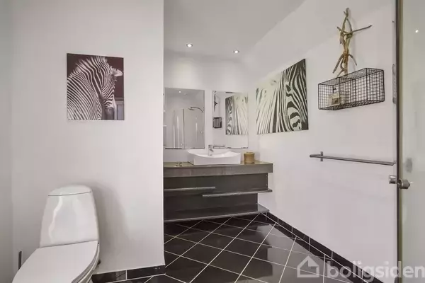 Et moderne badeværelse med en fritstående hvid toiletstol. Sort-hvide zebra-kunstværker pryder væggene. Et spejl og en væghængt vask ses over en klinkegulv med sorte fliser.