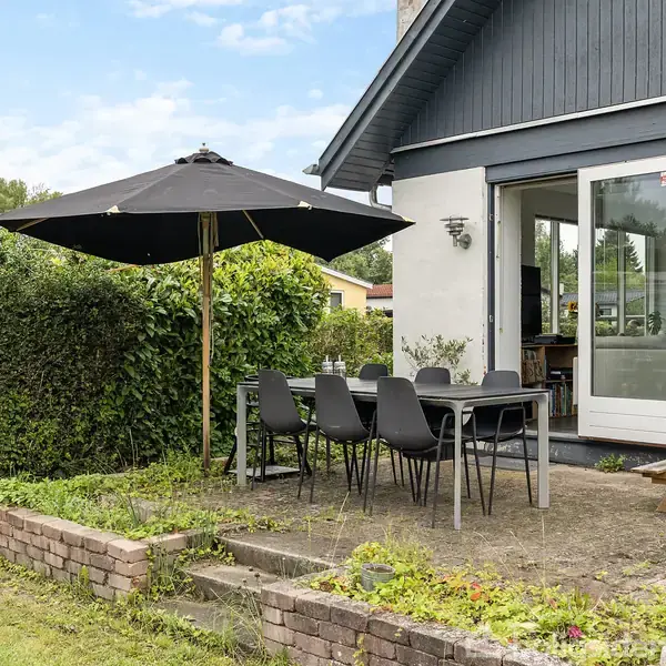 Terrasse med spisebord og seks sorte stole under en sort parasol. Området er omgivet af hække, og huset i baggrunden har store glasdøre, der fører ind til stuen.
