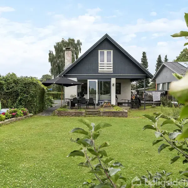 Et moderne hus med spidst tag står i forgrunden. Foran huset er der en terrasse med parasol og havemøbler. Omgivet af grønne plæner og buske i et beboelsesområde.