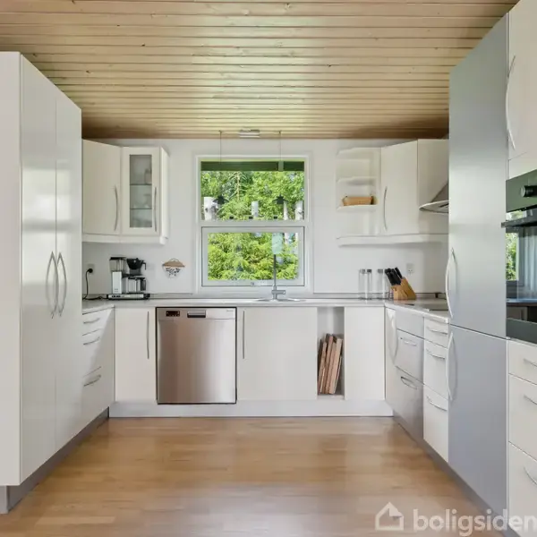 Hvidt køkken med moderne apparater som opvaskemaskine og indbygget ovn placeret i lyse træskabe. Store vinduer bagved viser frodigt grønt udendørs. Lyse trægulve og loft af træplanker.