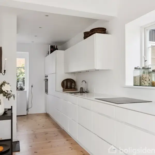 Et moderne køkken med hvide skabe strækker sig langs væggen. Glasbeholdere og dekorationer pynter overfladen. Trægulve og et stort vindue tilføjer lys, mens reoler rummer flasker og frugt.